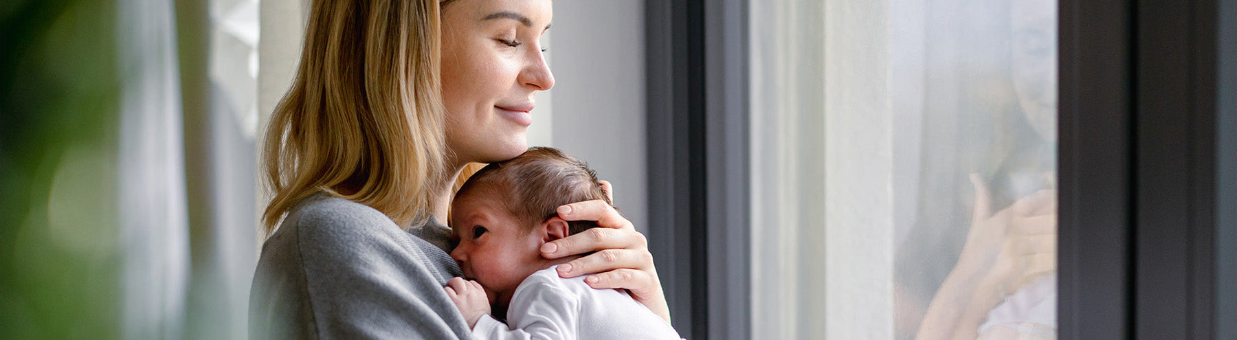 Mother holding her newborn baby close while standing by a window.