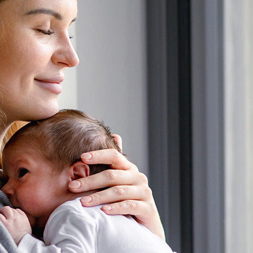 Mother holding her newborn baby close while standing by a window.