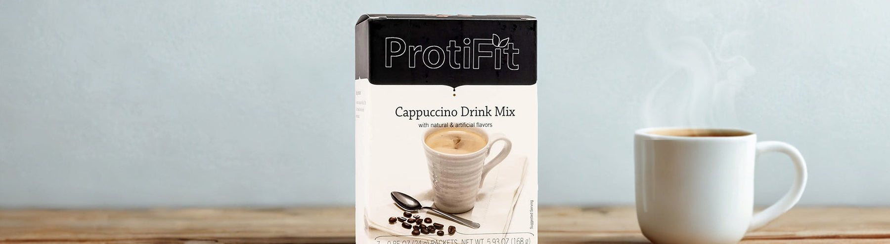Box of ProtiFit Protein Cappuccino Drink Mix displayed beside a steaming cup of it prepared on a wooden table.