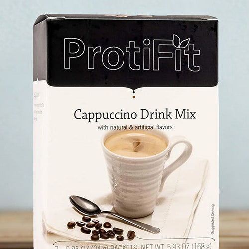 Box of ProtiFit Protein Cappuccino Drink Mix displayed beside a steaming cup of it prepared on a wooden table.