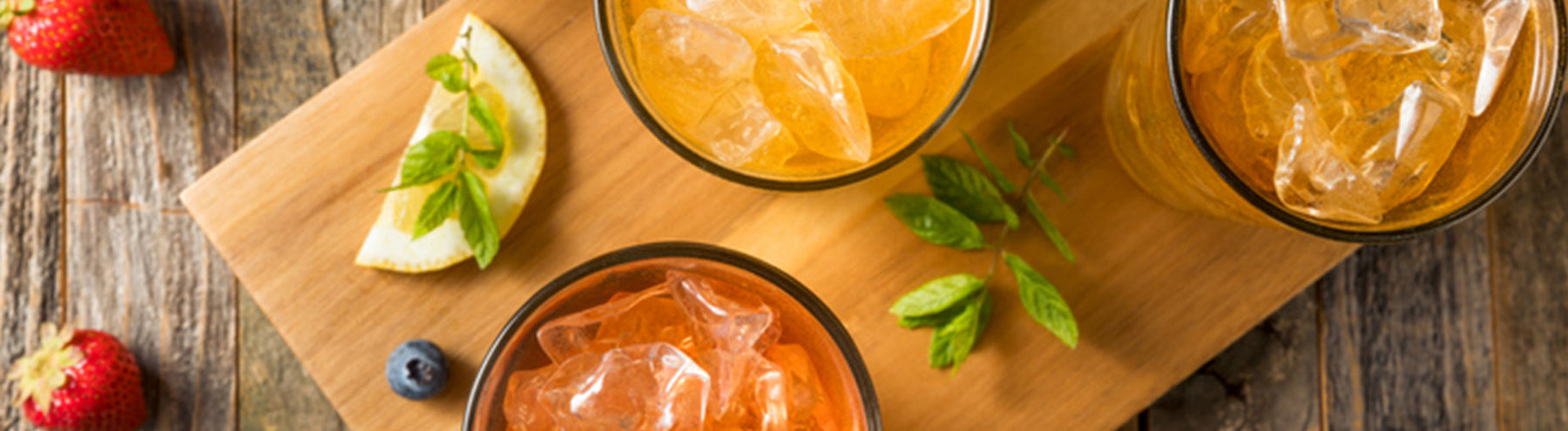 Iced fruit beverages with ice, lemon slices, berries, and fresh mint arranged on a wooden serving board.