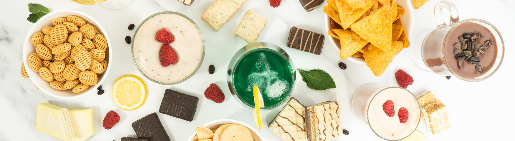 Overhead view of assorted snacks and desserts, including protein chips, wafers, chocolate pieces, protein pudding, protein shakes, lemon slices, raspberries, and a green beverage.