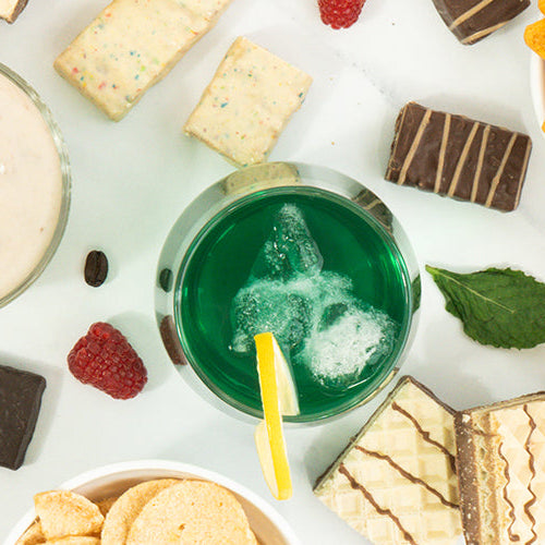 Overhead view of assorted snacks and desserts, including protein chips, wafers, chocolate pieces, protein pudding, protein shakes, lemon slices, raspberries, and a green beverage.