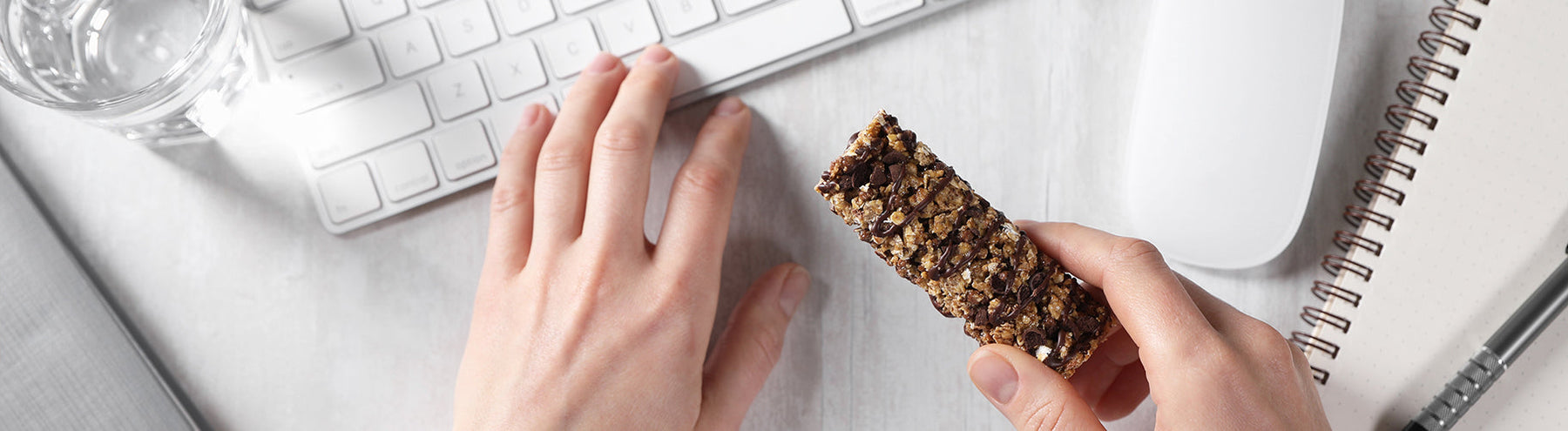 Person holding a granola-style protein bar while working at a computer with a notebook nearby.