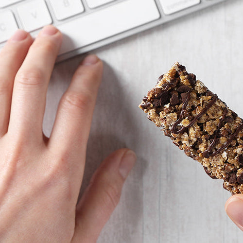 Person holding a granola-style protein bar while working at a computer with a notebook nearby.