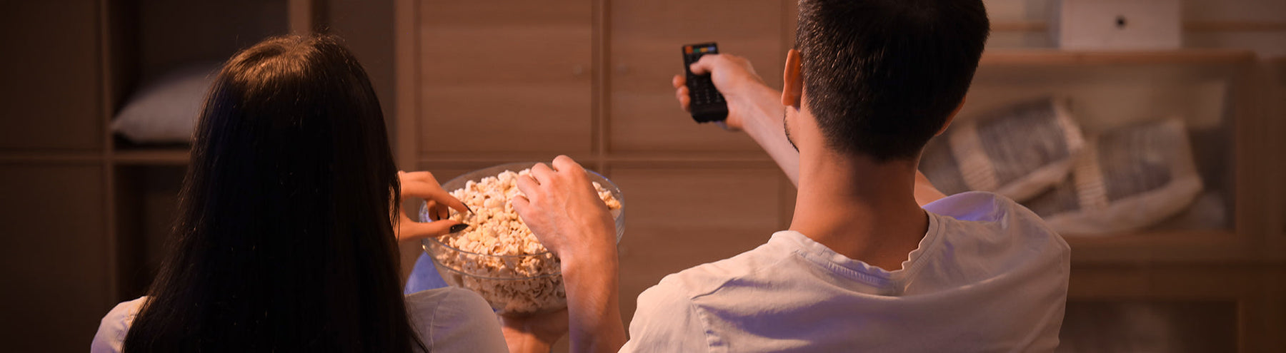 Couple sitting on the couch sharing a bowl of popcorn while one holds a TV remote during a movie night at home.