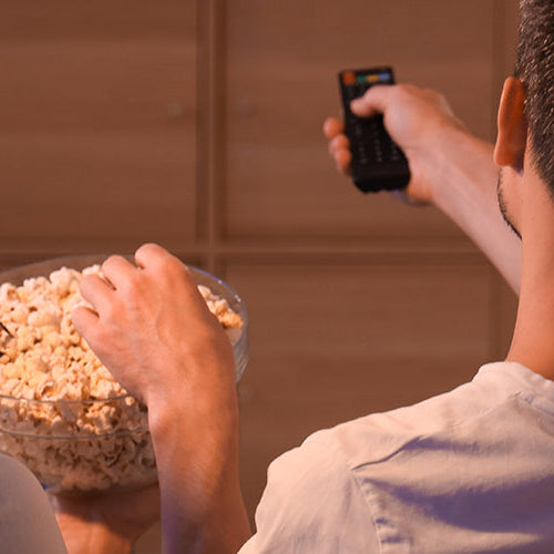 Couple sitting on the couch sharing a bowl of popcorn while one holds a TV remote during a movie night at home.